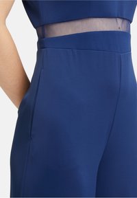 Mörkblå jumpsuit med ett transparent nätpanel vid midjan, mjukt tyg och sidofickor. Skräddarsydd passform med en ren design.