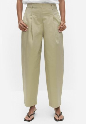 Vrouw draagt beige wijde broek met hoge taille en plooien, gecombineerd met zwarte teenslippers en een wit topje.