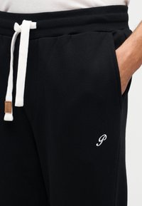 Μαύρο παντελόνι φόρμας από μαλακό ύφασμα με λευκό κορδόνι, πλαϊνές τσέπες και μικρό κεντημένο λογότυπο στο αριστερό μηρό.