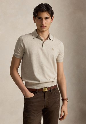 Jonge man met een beige polo met korte mouwen, een bruine broek, een bruine leren riem met gouden gesp en een bruine polshorloge, staand tegen een effen achtergrond.