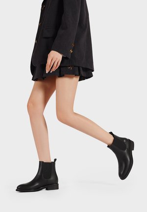 Stivaletti neri in pelle con finitura liscia, pannelli laterali elasticizzati, tacco basso e linguetta, abbinati a un blazer e una gonna neri.