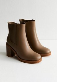 Brązowe skórzane botki za kostkę z ząbkowaną blokową obcasem i elastycznymi bocznymi panelami, umieszczone na białej powierzchni na tle jednolitego białego tła.