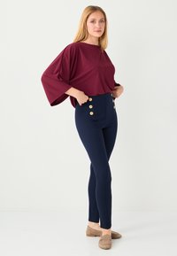Haut couleur marron style manches chauve-souris avec un col rond, associé à un legging taille haute bleu marine orné de boutons dorés. Tissu lisse et extensible.