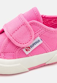 Růžová plátěná bota s suchým zipem, texturovaný svrchní materiál, bílá gumová podrážka a viditelné logo značky s červeným designem.