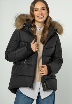 Schwarze Pufferjacke mit einer Kapuze aus Kunstfell, gestepptem Design, Reißverschlusstaschen und Strickbündchen über einem beigen Pullover und einem gestreiften Shirt.