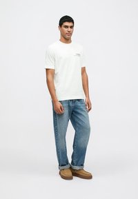 Tricou alb din bumbac cu mâneci scurte; blugi din denim albastru; pantofi maro din piele întoarsă; croială lejeră; design minimalist cu detaliu subtil de text.