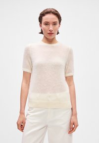 Svetlo béžový pletený sveter s krátkymi rukávmi, okrúhlym výstrihom a jemnou, chlpatou textúrou. Má o niečo tmavšiu farbu na leme.