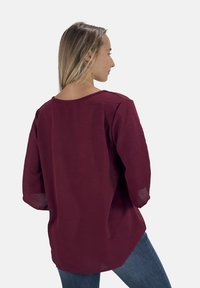 Blouse bordeaux à coupe ample, avec des manches larges et un col bateau. Tissu léger et texturé. Portée avec un jean, vue de dos.