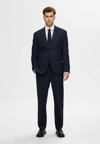 Marineblauer Anzug aus strukturiertem Stoff, einreihiger Blazer mit Revers, ergänzt durch ein weißes Hemd und eine dunkle Krawatte.
