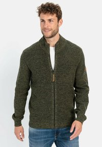 Dunkelgrüner gestrickter Reißverschluss-Cardigan mit hochgeschlossenem Kragen, strukturiertem Stoff und einem Reißverschluss mit einem Holzakzent. Darunter wird ein weißes T-Shirt getragen.