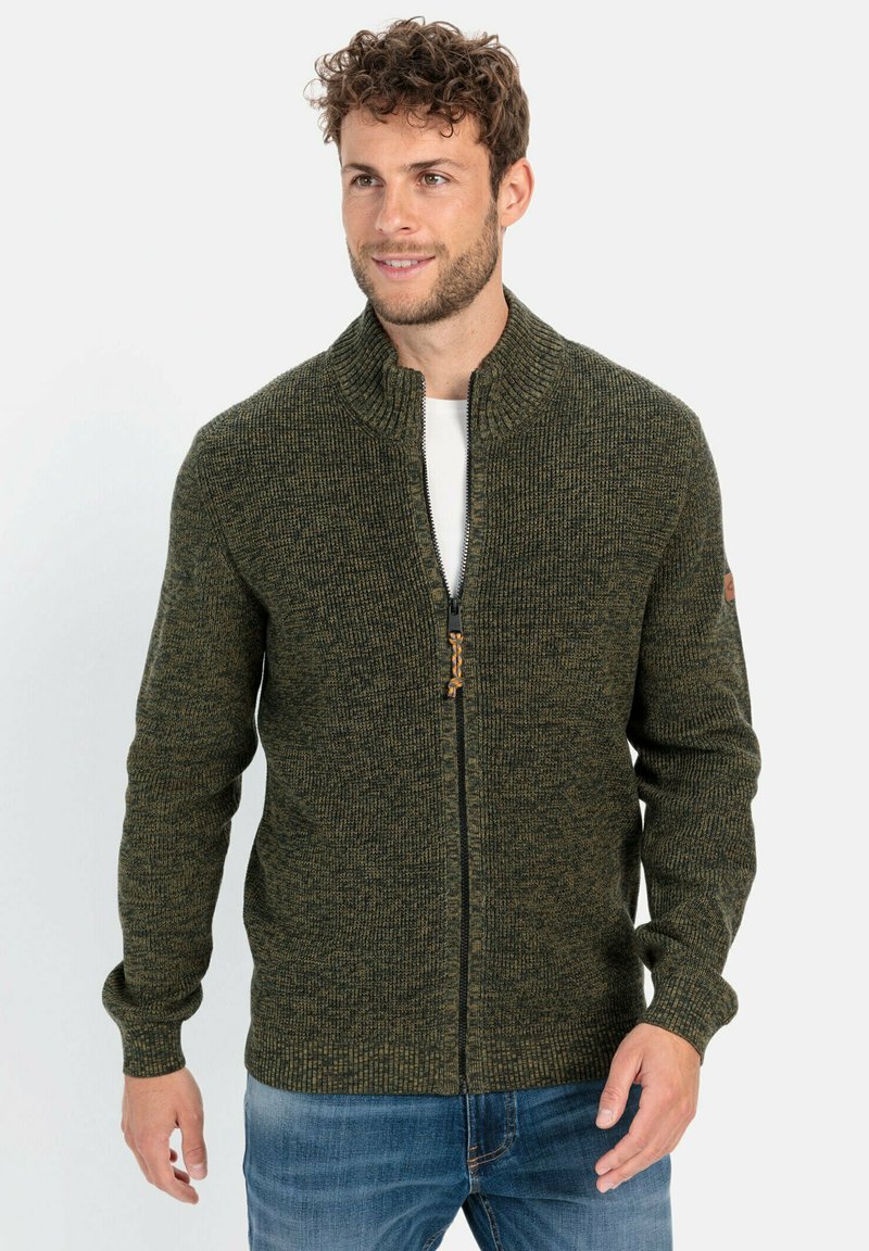 Dunkelgrüner gestrickter Reißverschluss-Cardigan mit hochgeschlossenem Kragen, strukturiertem Stoff und einem Reißverschluss mit einem Holzakzent. Darunter wird ein weißes T-Shirt getragen.
