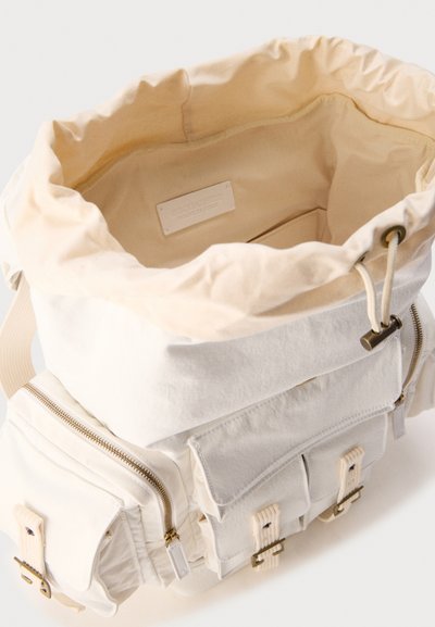 Σακίδιο πλάτης από καμβά σε κρεμ απόχρωση με ανοιχτό κορδόνι στην κορυφή, μπροστινές τσέπες με καπάκια και αγκράφες και πλαϊνές θήκες με φερμουάρ.