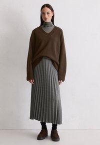 Bruine oversized trui met een V-hals, gelaagd over een grijze geribde coltrui. Grijze geplisseerde midi-rok en bruine instap schoenen.