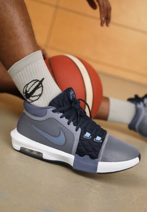 Zbliżenie osoby noszącej szare i czarne buty do koszykówki Nike oraz białe skarpety, trzymającej pomarańczową piłkę do koszykówki na podłodze hali gimnastycznej.