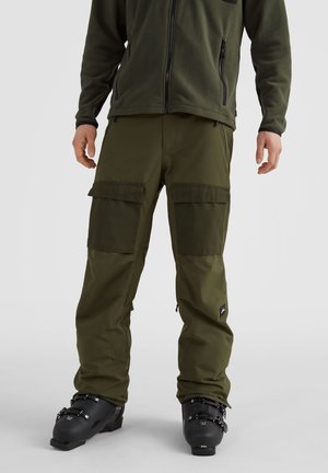 Mężczyzna w oliwkowozielonych spodniach bojowych i czarnych butach narciarskich, stojący na tle jasnego, jednolitego tła.
