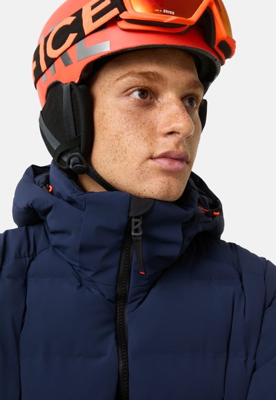 Navy padded jacket with high collar and zipper, featuring a logo detail. Accompanied by a bright orange helmet and goggles.