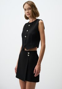 Top nero senza maniche con orlo a punta e bottoni a forma di fiocco argento, abbinato a una mini gonna nera coordinata con bottoni simili e design avvolgente.