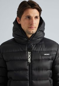 Sort pufferjakke med høj krave, horisontal quiltet design og frontlynlås. Logo lap på brystet og mærket lynlåslyft.
