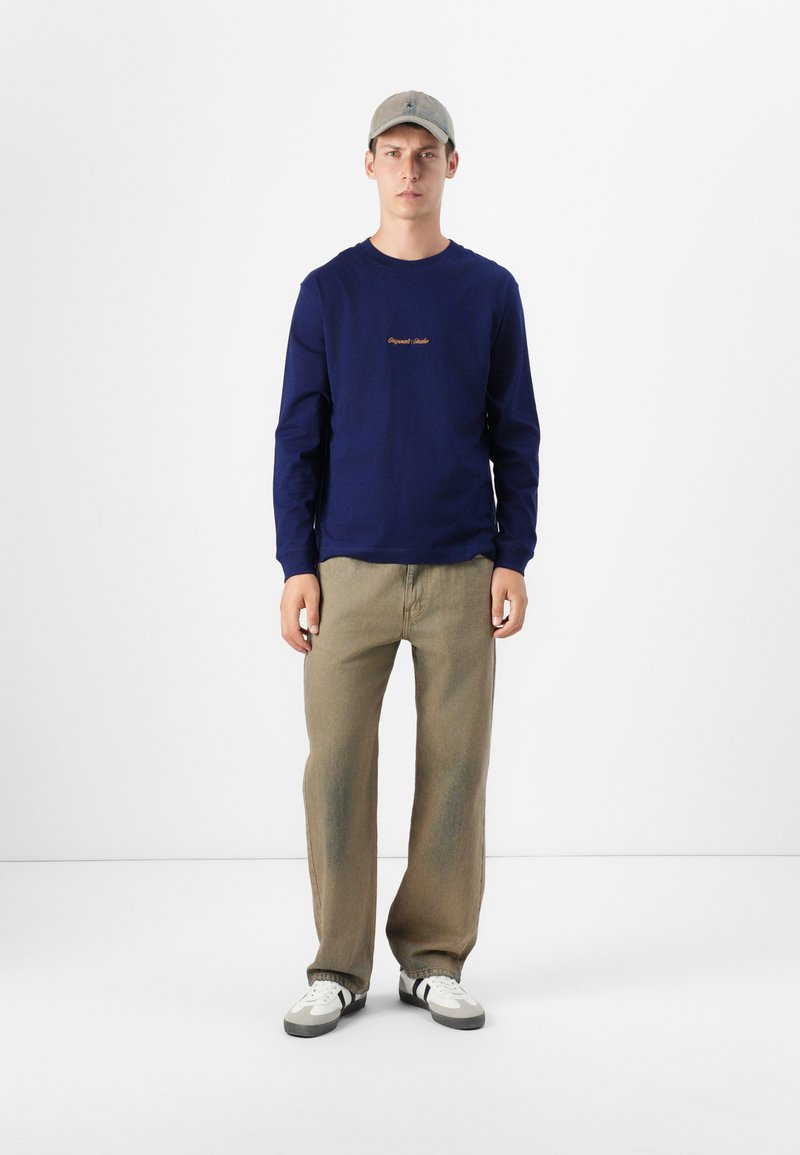 Młody mężczyzna stoi na białym tle, ubrany w granatową koszulę z długim rękawem, beżowe spodnie i szary kapelusz, z neutralnym wyrazem twarzy.