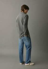Siva majica z dolgimi rokavi in sproščenim krojem, v kombinaciji z ohlapnimi modrimi jeans hlačami. Obleko dopolnjujejo svetle teniske. Nevtralna ozadje.