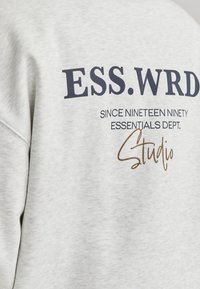 Gray sweatshirt with printed text in navy and brown. Features "ESS.WRD" and "Studio" with textured fabric and crewneck design.