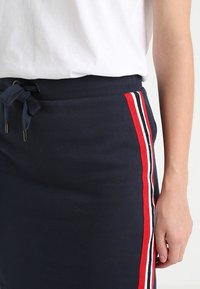 Primo piano di una gonna blu scuro con strisce laterali rosse, bianche e nere, vita a coste, nastri da stringere, abbinata a una maglietta bianca e con un braccio visibile.