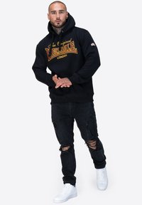 Felpa nera con grafica testuale arancione e gialla, abbinata a jeans neri scoloriti e sneakers bianche. Presenta una tasca a marsupio e cappuccio con cordino.