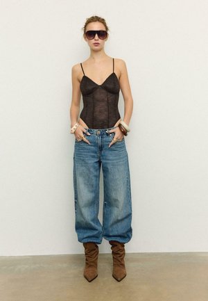 Vrouw die een zwart kanten bodysuit draagt, losse blauwe spijkerbroek, bruine slungelige laarzen, grote armbanden en oversized zonnebril, staand tegen een simpele muur.