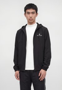 Junger Mann trägt eine schwarze Kapuzenjacke mit Reißverschluss und Logo, darunter ein weißes T-Shirt und schwarze Hose, steht vor einem schlichten Hintergrund.
