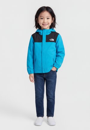 Ungt barn smilende, iført en blå og sort hættejakke, mørke jeans og hvide slip-on sko, stående mod en ensfarvet baggrund.