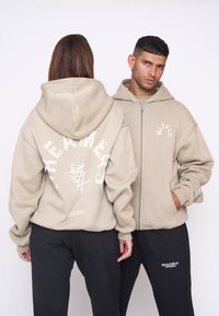 Beige zip-up hoodie lavet af blødt stof, med et stort hvidt grafisk design på ryggen og et mindre logo på forsiden. Sorte bukser med lommer.