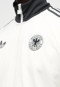 Nærbillede af en hvid sportjakke med sort krave og Adidas-logo, med Deutscher Fussball-Bund-emblemet på brystet.