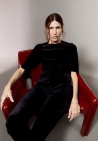 Zwarte fluwelen jumpsuit met korte mouwen, getailleerd ontwerp, gedragen terwijl zittend op een glanzende rode plastic stoel tegen een neutrale achtergrond.