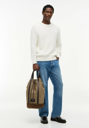 Uomo che indossa un maglione bianco a struttura, jeans blu, scarpe nere, che tiene una grande borsa a mano beige e marrone con manici scuri.