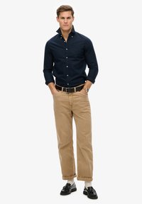Navy button-up shirt, khaki pants, black belt, black loafers with tassels, and white socks. Model stands in front of a plain white background.