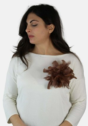 Kobieta o ciemnych włosach, ubrana w biały top ozdobiony dużym brązowym pióropuszem po lewej stronie, patrząca w dół i na prawo.