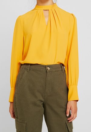 Blusa amarilla de manga larga con escote en forma de llave plisado combinada con pantalones cargo verde oliva de cintura alta y bolsillos laterales.