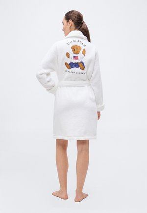 Woman with dark hair in ponytail wearing a white bathrobe with a teddy bear and "Polo Bear by Ralph Lauren" embroidered on the back.