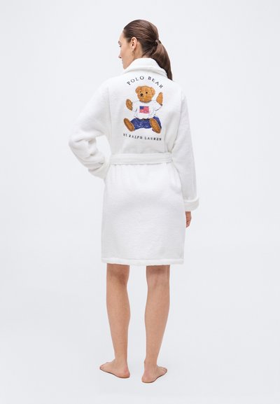 Kvinna med mörkt hår i hästsvans som bär en vit morgonrock med en teddybjörn och texten "Polo Bear by Ralph Lauren" broderad på ryggen.