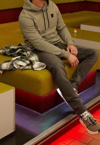 Sudadera verde claro con un logo, jeans grises y zapatillas de colores mixtos. Sentado en un banco amarillo en un interior moderno con iluminación ambiental.