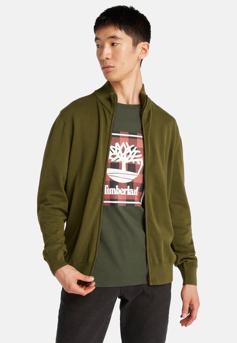 Olivgrön zip-up jacka med hög krage, ribbad mudd, lager över en mörkgrön T-shirt med en vit logotyp och rutigt mönster.