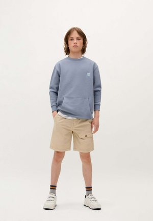 Sudadera azul con bolsillo, pantalones cortos beige con bolsillo lateral tipo cargo y zapatillas blancas. El conjunto tiene un ajuste relajado y un aspecto en capas.