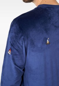Tumši zils samta sporta krekls ar gludu tekstūru, ar izšūtiem dizainiem uz piedurknēm, kas attēlo liesmas un ugunskuru.