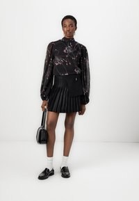Blusa nera con motivo floreale e maniche a sbuffo lunghe e trasparenti, abbinata a una gonna nera plissettata e mocassini neri. Tra gli accessori, una borsa nera.