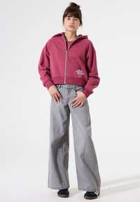 Sudadera con cremallera, recortada, en color burdeos con el texto "HAZTE ORGULLOSO"; combinada con vaqueros de pierna ancha en gris claro y zapatillas negras.