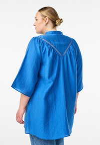 Blue textured blouse with a v-shaped back yoke, short sleeves, and decorative stitching along the yoke. Casual, relaxed fit.