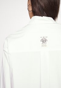 Vit skjorta med knapp-up och en baketikett som har en logotyp och text som firar 50 år. Slät tygkvalitet, klassisk krage och avslappnad passform.