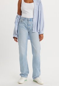 Personne portant un jean bleu clair, un débardeur blanc, une chemise rayée bleue portée ouverte et des baskets blanches avec des touches vertes sur la semelle.
