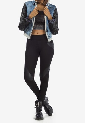 Femme portant une veste en denim et cuir noir, un crop top noir, des leggings noirs avec des patchs, et des bottes de combat noires cloutées, debout les jambes croisées.