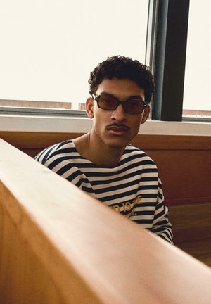 Jeune homme aux cheveux bouclés et moustache portant des lunettes teintées et une chemise rayée noir et blanc, assis près d'une fenêtre dans une cabine en bois.